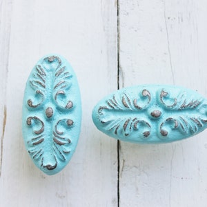 May include: Two oval-shaped cabinet knobs with a distressed light blue finish and intricate silver-grey floral designs.