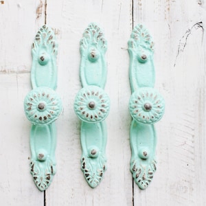 May include: Three antique-style cabinet knobs with a distressed, light blue finish and silver accents. The knobs have a floral design and are mounted on a white wooden surface.