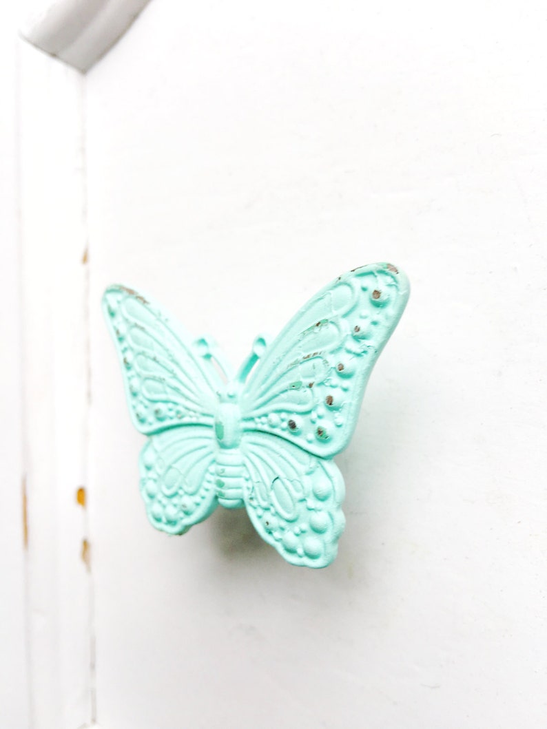 K&ouml;nnte beinhalten: Ein t&uuml;rkisblauer Schrankknauf in Schmetterlingsform mit aufwendigen Pr&auml;gungen. Der Knauf ist auf einer wei&szlig;en Oberfl&auml;che montiert, was sein filigranes Design und seine Farbe hervorhebt. Die Fl&uuml;gel des Schmetterlings sind ausgebreitet und verleihen der Dekoration einen Hauch von Natur.