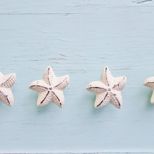 May include: Four white starfish-shaped decorative objects with dark accents are arranged horizontally on a light blue wooden surface. The starfish have a textured surface and are evenly spaced, creating a coastal or nautical theme.