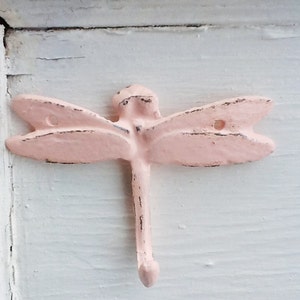 May include: A pink metal dragonfly hook. The hook is in the shape of a dragonfly with wings and a long tail that ends in a hook.