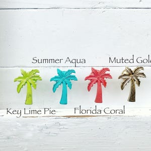 May include: Five palm tree shaped metal hooks in different colors: muted gold, summer aqua, key lime pie, florida coral, and a bright green. The hooks are mounted on a white painted wooden surface.