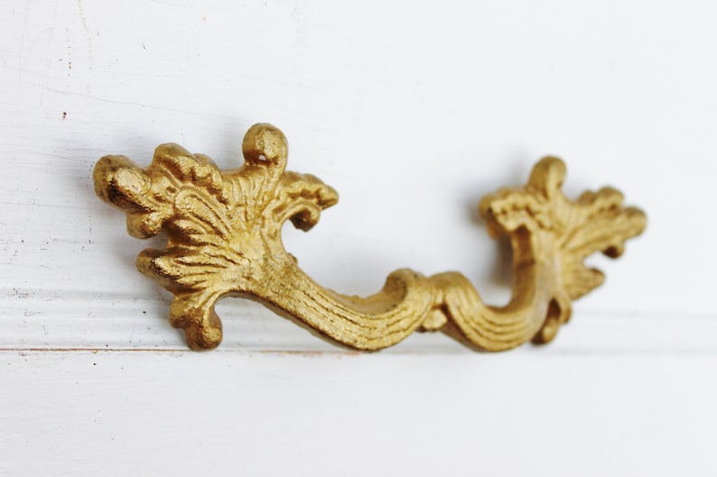 May include: Gold-toned ornate drawer pull with a decorative scroll design. The pull has a floral design on each end.