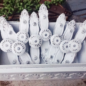 K&ouml;nnte beinhalten: Eine wei&szlig;e Holzkiste gef&uuml;llt mit wei&szlig;en Metall-Schubladengriffen mit verzierten Designs. Die Kiste hat ein dekoratives geschnitztes Design auf der Vorderseite.