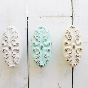 May include: Three oval-shaped cabinet knobs with a distressed white, light blue, and cream finish. Each knob features a raised, ornate design.