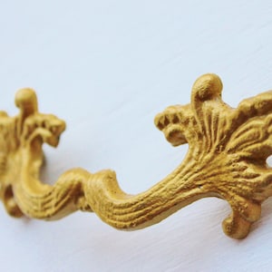 May include: A gold-colored metal drawer pull with a decorative, ornate design. The pull has a curved shape with intricate details resembling leaves and swirls.