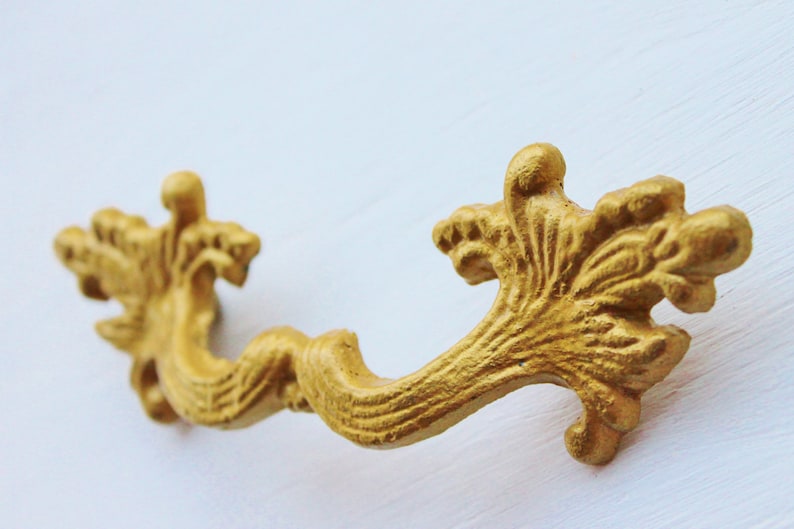 May include: A gold-colored metal drawer pull with a decorative, ornate design. The pull has a curved shape with intricate details resembling leaves and swirls.