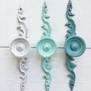 May include: Three decorative cabinet knobs with ornate scrollwork designs. The knobs are painted in shades of white, light blue, and teal.