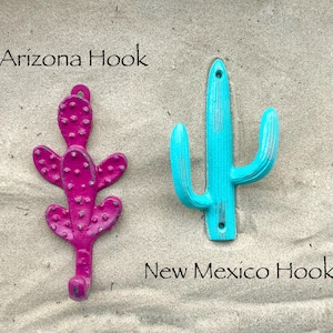 May include: Two metal cactus-shaped wall hooks. One is magenta with a textured surface and the text "Arizona Hook". The other is turquoise with the text "New Mexico Hook". Both hooks have a hole for hanging.