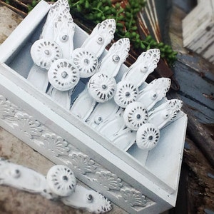 K&ouml;nnte beinhalten: Eine wei&szlig;e Holzkiste gef&uuml;llt mit wei&szlig;en Keramik-Schubladengriffen mit verzierten Designs. Die Griffe haben eine silberne Oberfl&auml;che und sind in einer Reihe angeordnet.