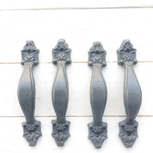May include: Four gray cast iron drawer pulls with ornate designs. The pulls are arranged in a row on a white wooden surface.
