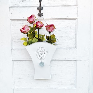 May include: A white metal planter with a decorative design hangs from a hook on a white wooden door. The planter is filled with pink roses.