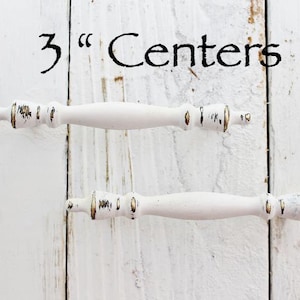 May include: Two white cabinet pulls with gold accents. The pulls are 3 inches apart. The text "3" Centers" is written above the pulls.
