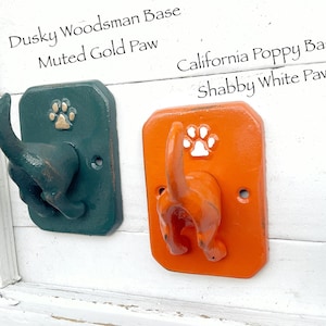 May include: Two decorative wall hooks shaped like dog tails. One is dark green with a gold paw print, the other is orange with a white paw print. The text on the top says "Dusky Woodsman Base, Muted Gold Paw" and "California Poppy Base, Shabby White Paw".