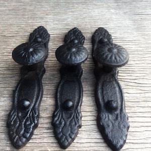 May include: Three black cast iron drawer pulls with ornate designs. Each pull has a round knob with a raised center and a decorative base.