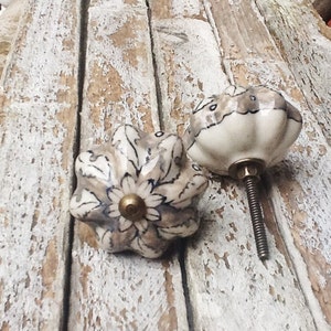 May include: Two ceramic cabinet knobs with a floral design in shades of gray and white. The knobs have a brass screw and are shown on a weathered wooden surface.
