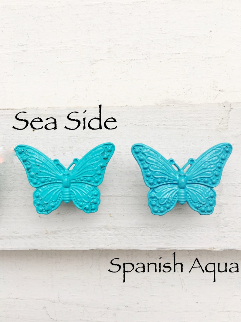 以下が含まれることがあります： 「Sea Side」と「Spanish Aqua」という文字が書かれた、ターコイズブルーの蝶の形をした金属製の引き出しノブが2つあります。
