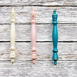 May include: Three decorative cabinet knobs with gold accents. The knobs are painted in white, pink, and teal.