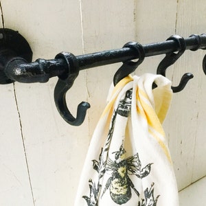 May include: Black metal pipe towel rack with four hooks, mounted on a white wall. A white tea towel with a yellow stripe and a black bee design hangs on one of the hooks.