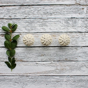 May include: Three white ceramic knobs with a floral design, arranged in a row on a whitewashed wooden surface. A green leafy branch is to the left of the knobs.