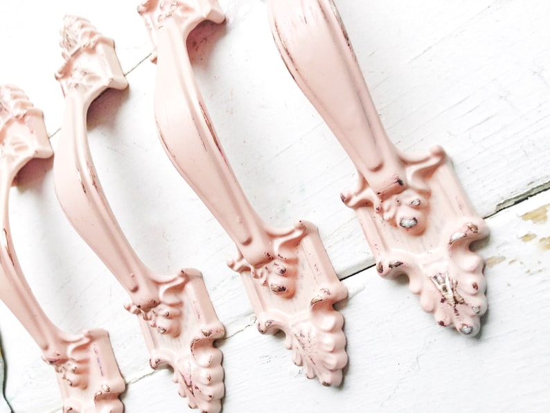 May include: Four pink metal drawer pulls with ornate designs. The pulls have a distressed finish and are perfect for adding a vintage touch to furniture.