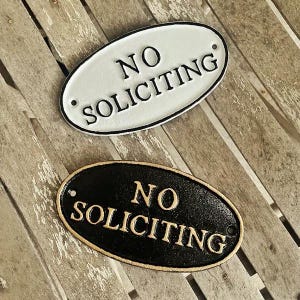 May include: Two oval-shaped signs with the text "No Soliciting" in black lettering. The top sign is white with black lettering and the bottom sign is black with gold lettering.