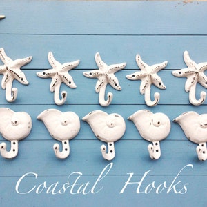 May include: White ceramic hooks shaped like seashells with starfish details. The hooks are mounted on a light blue wooden surface. The text "Coastal Hooks" is written below the hooks.