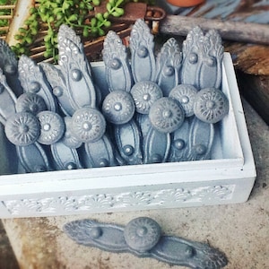 May include: A white wooden box filled with gray metal drawer pulls. The box has a decorative floral design on the front. The drawer pulls have a rounded, ornate design.