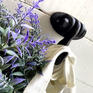 May include: A black, rustic metal hook with a curved design, mounted on a white wooden surface. A cream-colored cloth is hanging from the hook. Purple lavender and green foliage are in the background.
