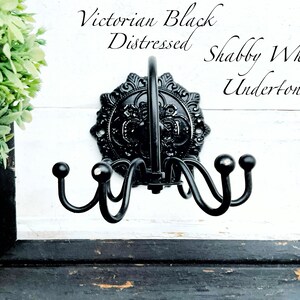 May include: Black metal coat rack with ornate details and a distressed finish. The rack has four hooks and is mounted on a white wall with the text "Victorian Black Distressed Shabby White Undertones".