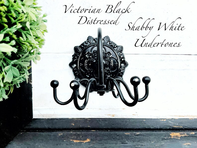 May include: Black metal coat rack with ornate details and a distressed finish. The rack has four hooks and is mounted on a white wall with the text "Victorian Black Distressed Shabby White Undertones".