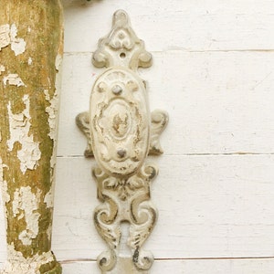 May include: White painted metal door knob with an ornate design. The knob has a keyhole and is attached to a white painted wooden surface.