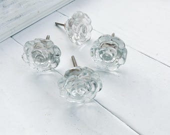 May include: Four clear glass rose-shaped cabinet knobs with silver stems. The knobs are on a white, textured surface. The floral design adds a decorative touch to furniture. The knobs are a decorative item.