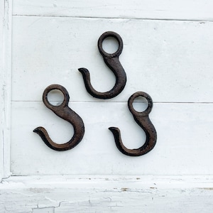 May include: Three rustic black metal hooks with a rounded top loop. The hooks are attached to a white painted wooden surface.
