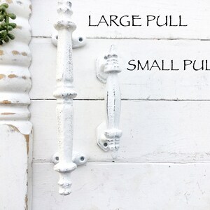 May include: Two white, distressed-finish pull handles, one large and one small, are displayed against a white, wooden background. The text "LARGE PULL" and "SMALL PULL" are above the respective handles. The handles are likely for furniture.