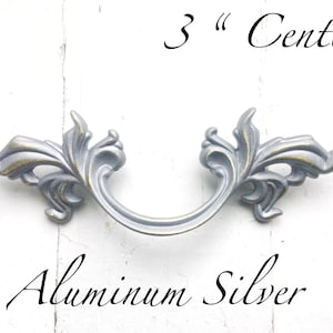 May include: Silver-colored cabinet pull with an ornate, floral design. The handle is 3 inches (7.6 cm) between the centers of the mounting holes. The text "Aluminum Silver" is below the handle.
