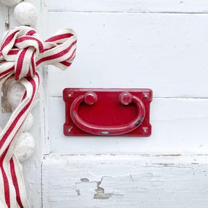 May include: A red metal drawer pull with a rounded handle, mounted on a white painted wooden surface. A red and white striped ribbon is tied in a bow on the left side of the image.