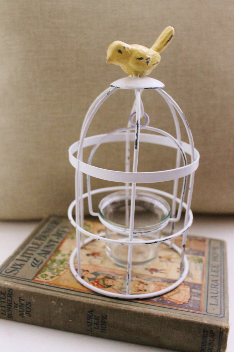May include: White metal birdcage with a yellow bird on top. The cage has a glass votive holder inside. The cage is sitting on a vintage book.