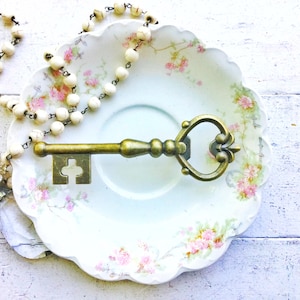 May include: A vintage-style key-shaped bottle opener with a heart-shaped opening, resting on a white porcelain plate with a floral pattern. A white beaded necklace with a rhinestone brooch is also on the plate.