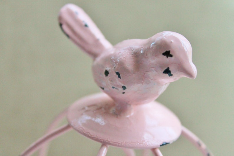 May include: A pink bird figurine with black accents sits atop a pink metal birdcage. The birdcage has a round base and multiple metal bars.