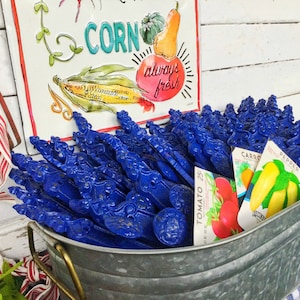 May include: A galvanized metal bucket filled with blue metal plant markers. The markers have a decorative design and are arranged in a pile.  The bucket is sitting on a blue and white checkered tablecloth.  There are three seed packets visible in the bucket, labeled "Tomato 25c", "Carrot", and "Pepper".