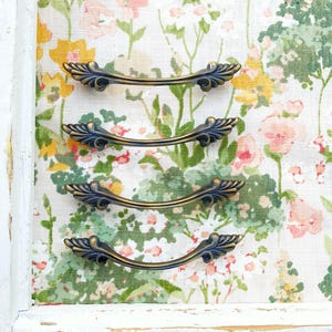 May include: Four antique bronze drawer pulls with a decorative leaf design. The pulls are curved and have a dark blue finish with gold accents. They are set against a floral background with pink, yellow, and green flowers.