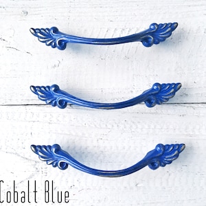 May include: Three cobalt blue cabinet pulls with a decorative, ornate design. The pulls have a curved shape with detailed, leaf-like accents on each end. The text "Cobalt Blue" is at the bottom of the image.