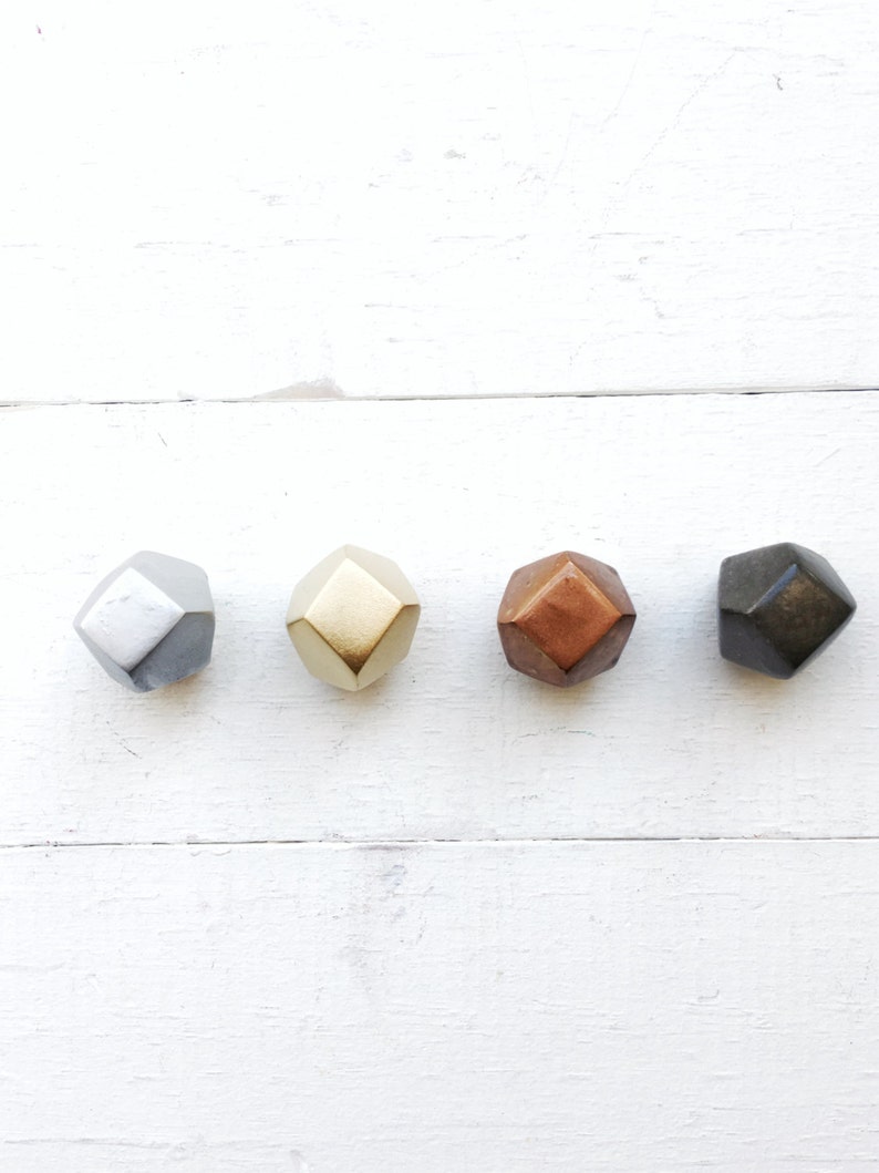 May include: Four geometric shapes in different metallic colors: silver, gold, copper, and black. The shapes are arranged in a row on a white wooden surface.