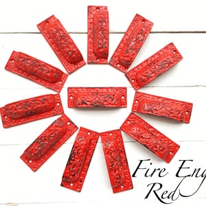 May include: Twelve red metal drawer pulls with an ornate design. The pulls are arranged in a circle on a white wooden surface. The text "Fire Engine Red" is written below the pulls.