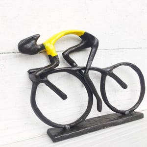 May include: Black metal sculpture of a cyclist in a yellow jersey riding a bicycle with black wheels. The sculpture is mounted on a black metal base.