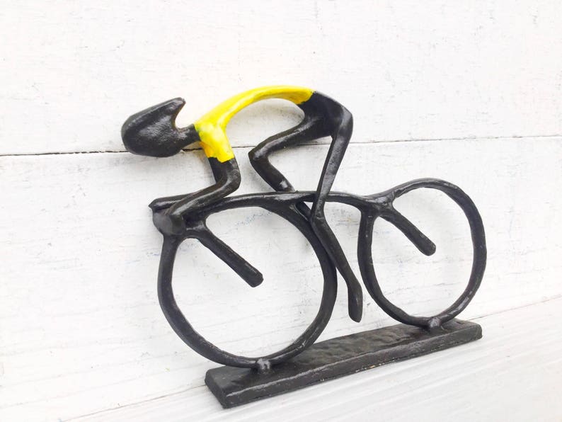 May include: Black metal sculpture of a cyclist in a yellow jersey riding a bicycle with black wheels. The sculpture is mounted on a black metal base.