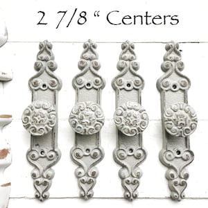May include: Four antique-style door knobs with ornate details in a light gray finish. The knobs are mounted on a white background and have a 2 7/8" center to center spacing.