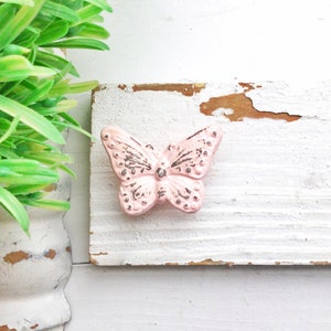 Peut inclure: Un bouton en c&eacute;ramique en forme de papillon rose avec des accents argent&eacute;s sur une surface en bois peinte en blanc.