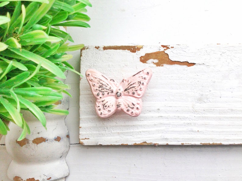 Peut inclure: Un bouton en c&eacute;ramique en forme de papillon rose avec des accents argent&eacute;s sur une surface en bois peinte en blanc.